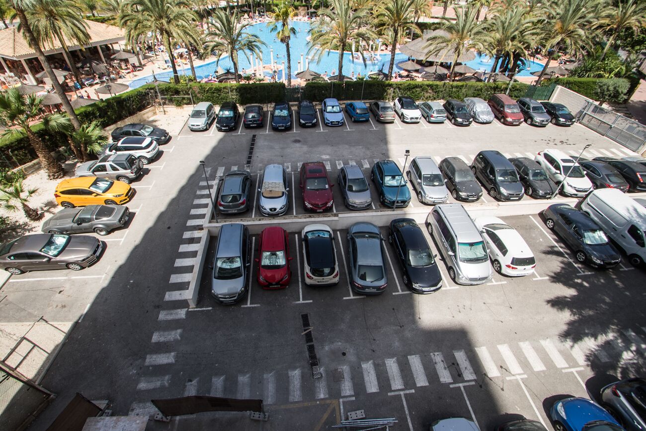 Un estacionamiento con varias filas de coches aparcados, un paso de peatones y una zona de piscina con palmeras al fondo.