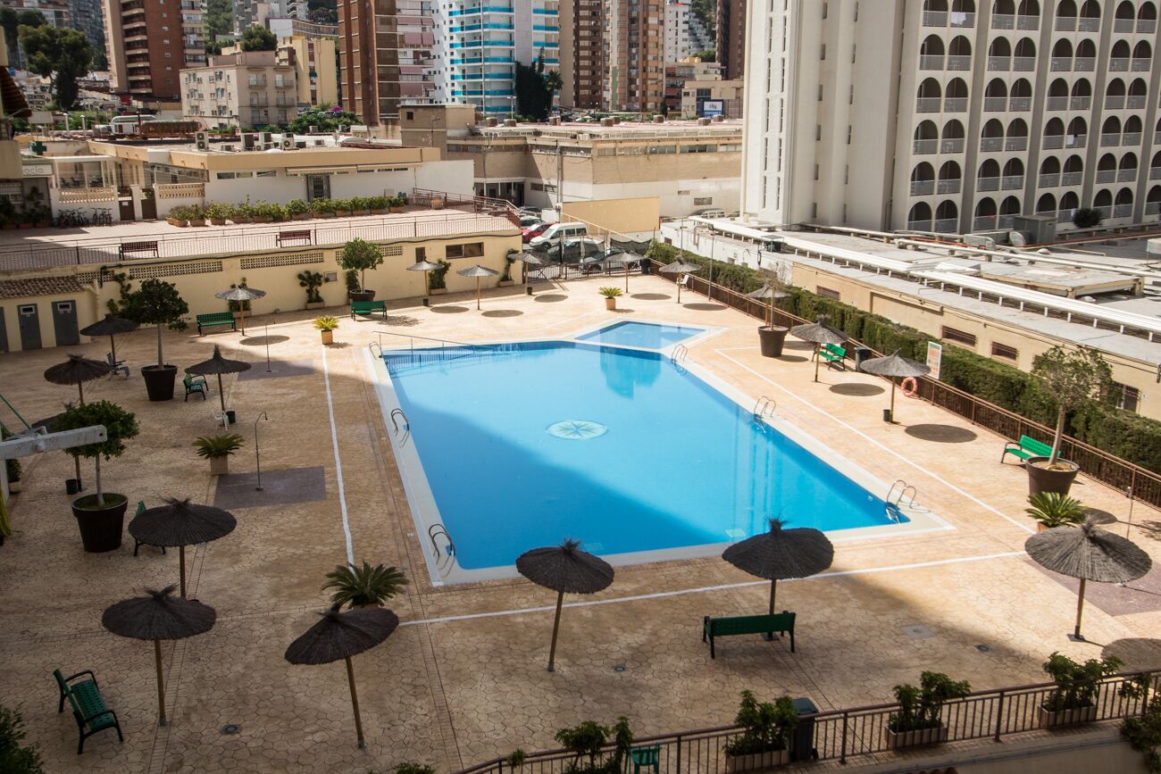 Un área de piscina en la azotea con piscina azul, rodeada de tumbonas y sombrillas, con edificios urbanos al fondo.