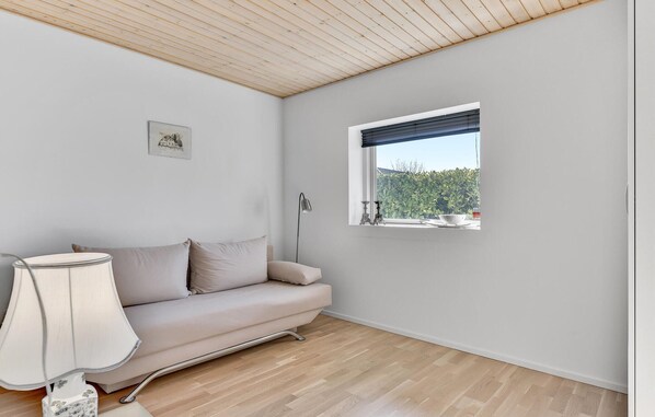 Quarto com teto de madeira, sofá bege, abajur, janela com vista para vegetação e um quadro na parede.