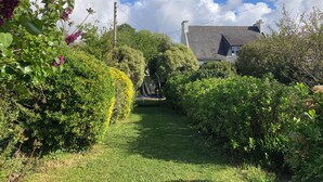 A garden pathway leading to a house with a sloped roof, surrounded by lush greenery and flowering plants. - Maison Familiale 7ch au Coeur de Ste Marine. Plage à 10m à Pieds ou 5 en Vélos ! (Combrit)