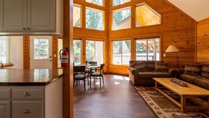 A spacious log cabin interior with a dining area, a living room with a sofa and a coffee table, and large windows allowing natural light. - Southgate Getaway of Yosemite (Fish Camp)
