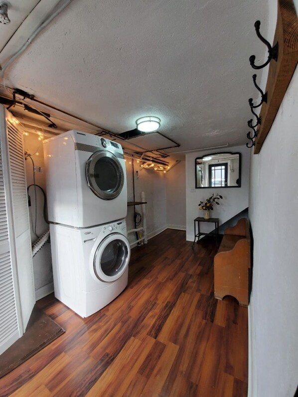 A compact laundry room with a stacked washer and dryer, wooden flooring, and a small seating area with a plant. - Private, Fully Renovated 1 bedroom Carriage House (Phoenixville)