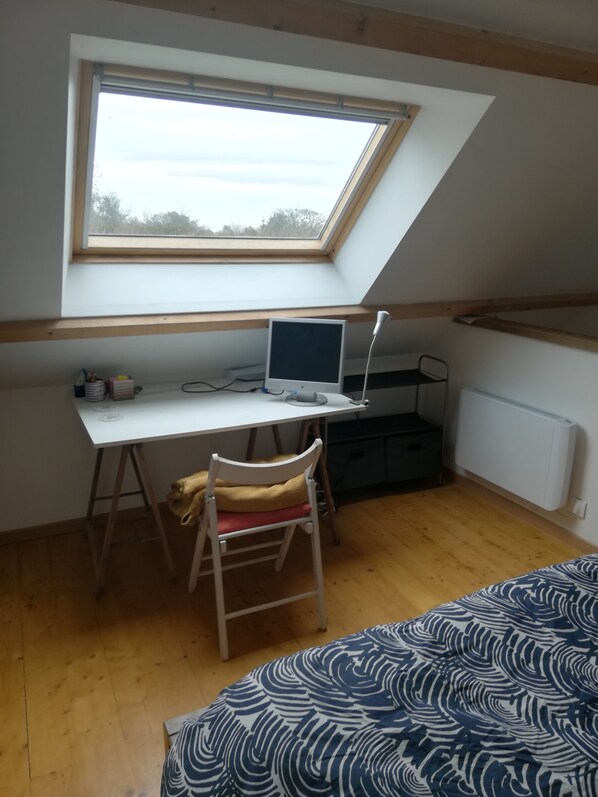 A small, single-room apartment with a sloped ceiling, a skylight, a white desk with a computer, a chair, a radiator, and a bed with a patterned bedspread.