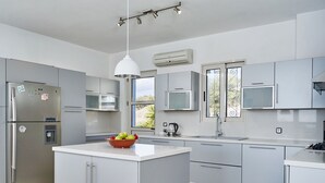 A modern kitchen with grey cabinets, a central island with a fruit bowl, stainless steel appliances, and a window with a view. - Luxury on Rhodes (Lardos)