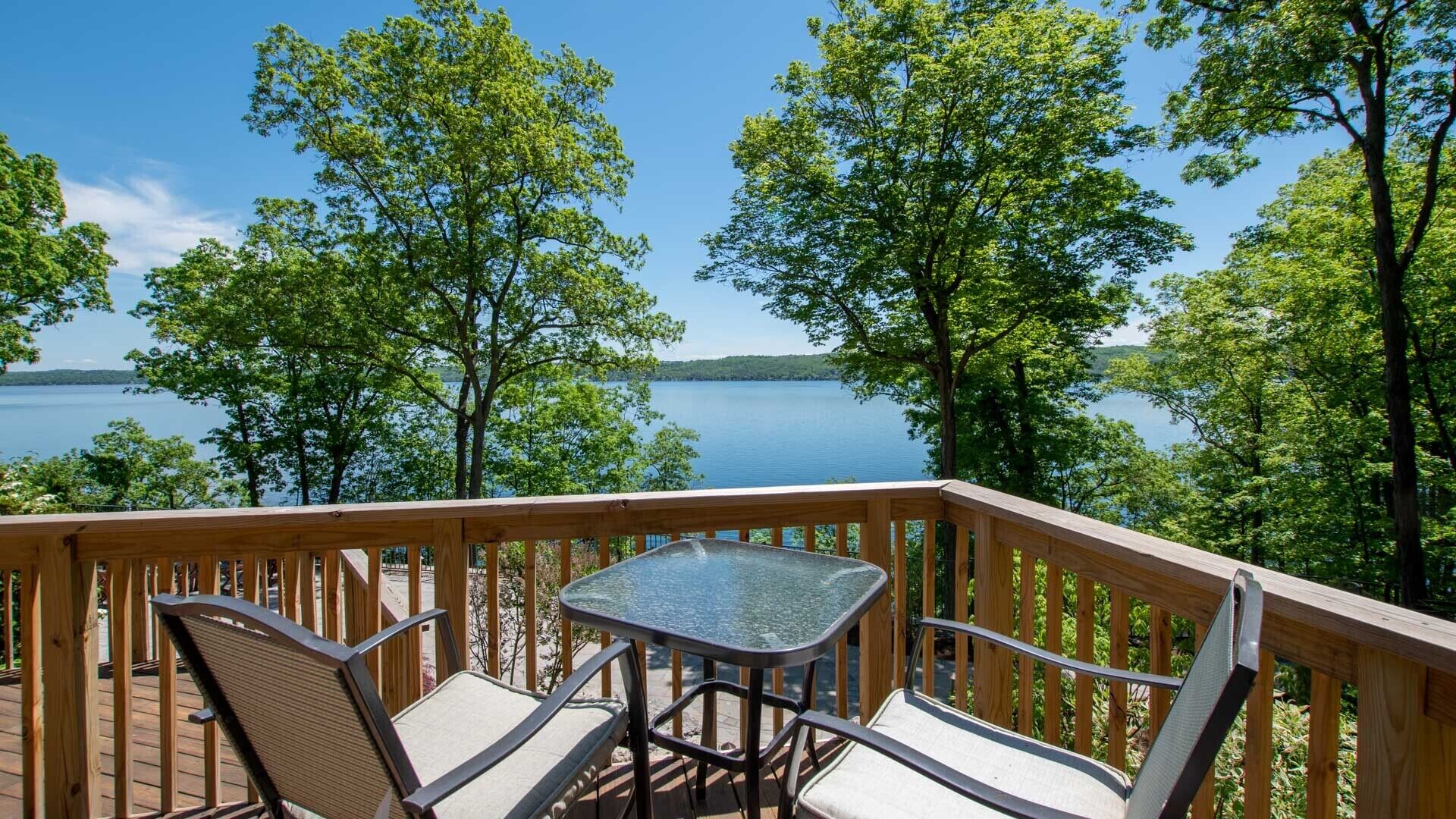 Une terrasse en bois avec une table en verre et deux chaises, donnant sur un lac bordé d’arbres.