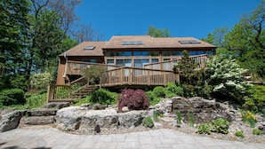 Une maison en bois à deux étages, dotée d’une terrasse et entourée d’une végétation luxuriante, avec un chemin de pierre.