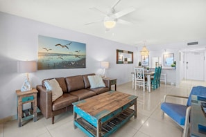 A living room with a brown leather sofa, a wooden coffee table, a dining area with a white table and chairs, and a ceiling fan. - Wake Up to Waves: Penthouse Condo w/ Direct Ocean Views & Heated Pool 🌊 (Satellite Beach)