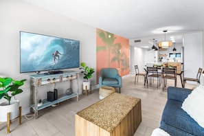 A modern living room with a flat-screen TV, a blue sofa, a wooden coffee table, and a dining area with chairs and a table.