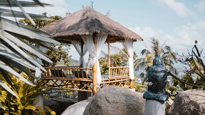 A thatched-roof gazebo with a wooden bench, surrounded by tropical foliage and large rocks. - Paradise Villa Eden (Koh Samui)