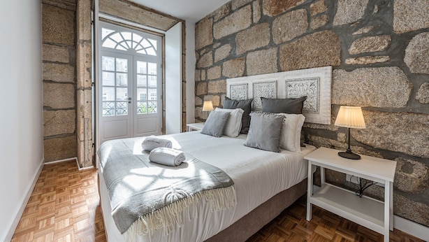 A bedroom with a stone wall, a bed with pillows, a bedside table with a lamp, and a door leading to an outdoor area. - GuestReady - Grandeur River Balcony Reborn (Porto)