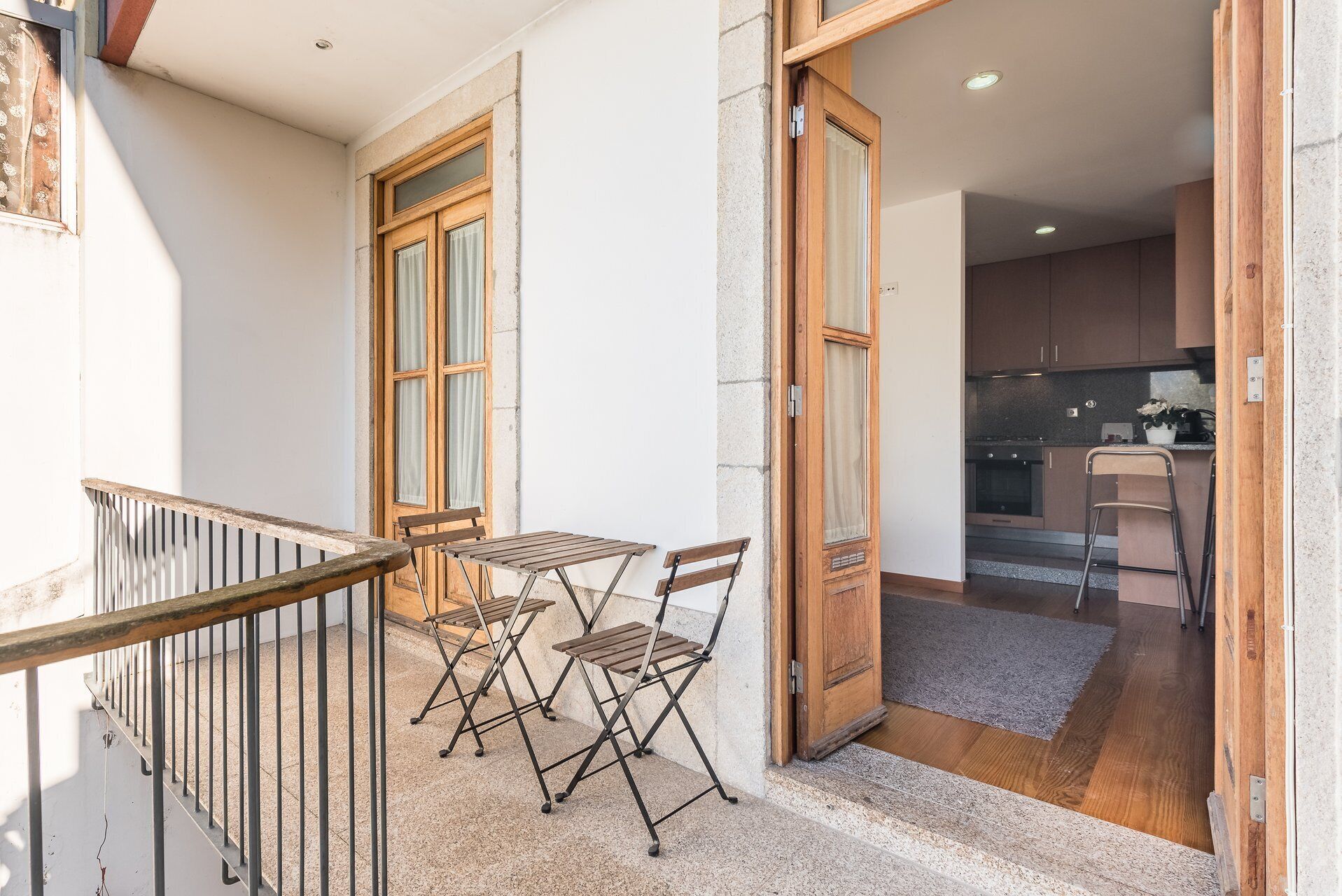 A balcony with a table and chairs, a wooden door leading to a kitchen, and a metal railing.