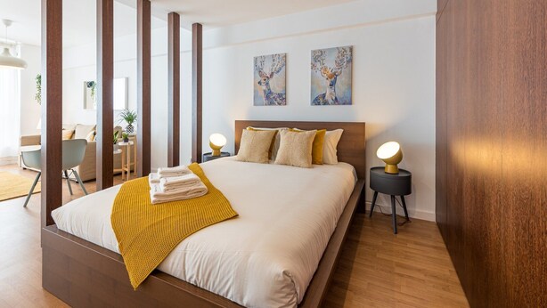 A modern hotel room with a large bed, two bedside lamps, a wooden headboard, and two abstract paintings on the wall. - GuestReady - Hemisphere Apt (Porto)