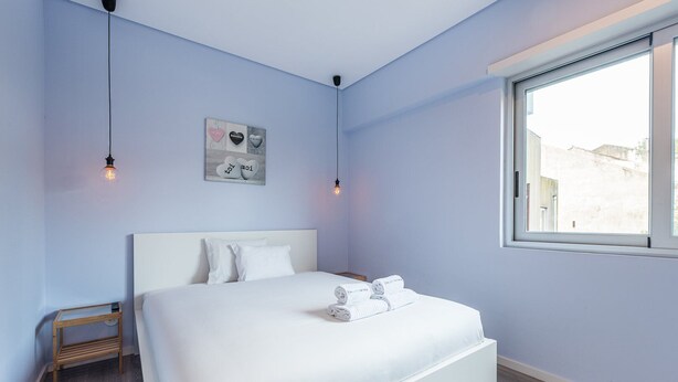 Une chambre avec un lit blanc, une table de chevet, un tableau décoratif au mur et une fenêtre.