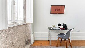 A hotel room with a white wall, a framed 'GUEST READY' sign, a small table with a coffee maker, a chair, and a window with white curtains.