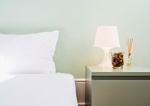 Una cama bien tendida con sábanas blancas, una lámpara de noche y un recipiente de vidrio con flores secas y difusores de aroma sobre una mesita.