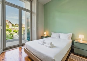 A hotel room with a bed, two bedside tables with lamps, a view through large windows, and a green wall.