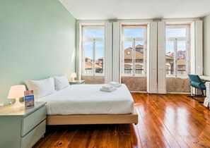 A hotel room with a bed, a desk, and a view of buildings through large windows.