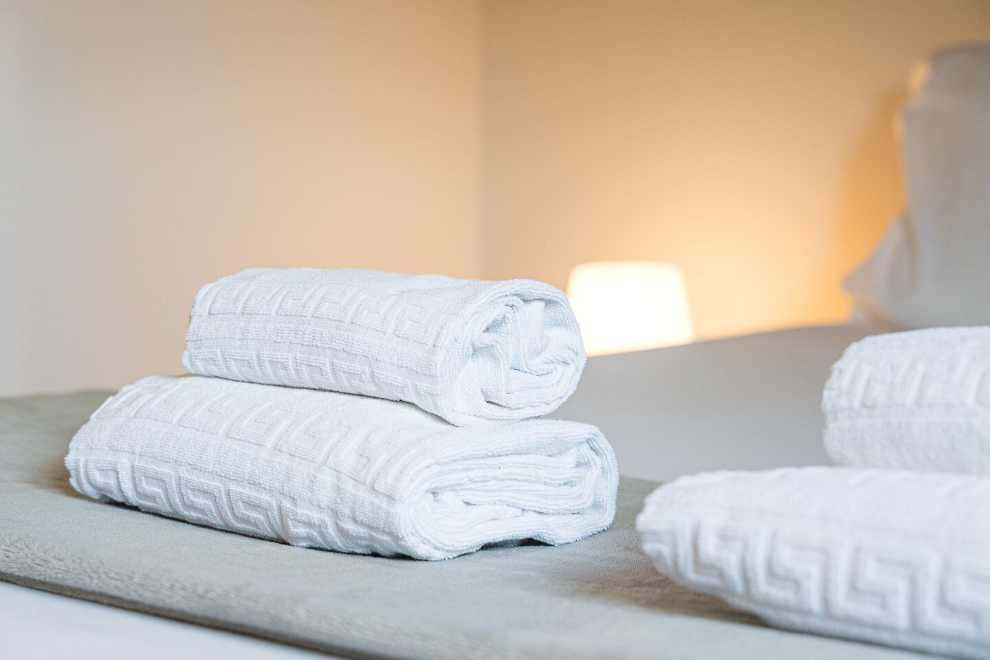 Toalhas brancas bem dobradas empilhadas sobre uma superfície texturizada, possivelmente uma cama ou porta-toalhas, em um ambiente com cores neutras e iluminação suave.