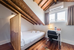 A bedroom with a bed, a desk, and a chair. There is a window with curtains, an air conditioning unit, and a wooden ceiling.