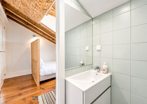 A bedroom with a wooden ceiling, a bed, and a bathroom with a white sink and green tiles.