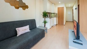 Uma sala de estar moderna com um sofá cinzento, uma mesinha, uma televisão e uma área de cozinha ao fundo.