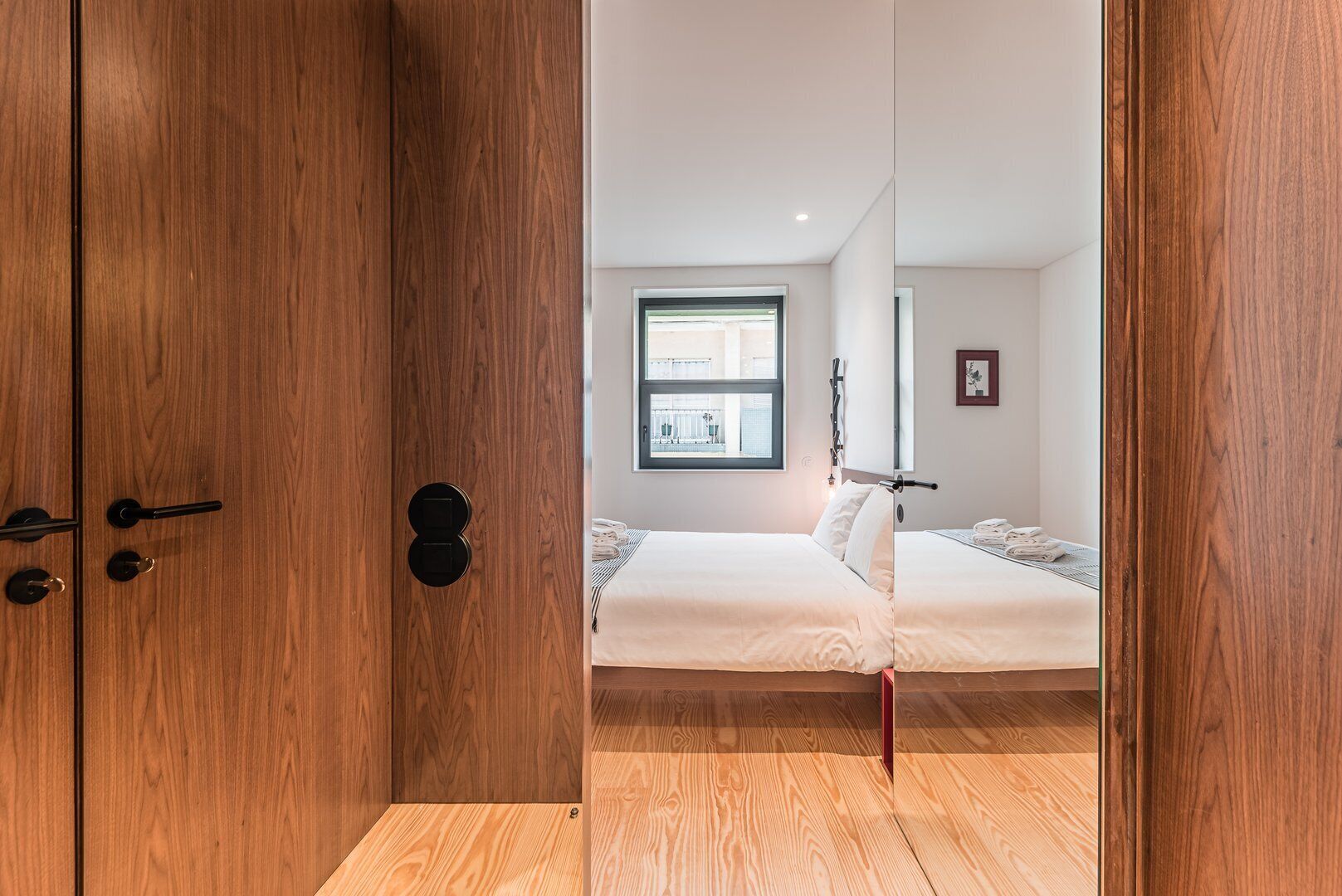 Quarto com piso de madeira, cama com roupa de cama branca, janela com vista e porta com puxador.