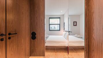 Quarto com piso de madeira, cama com roupa de cama branca, janela com vista e porta com puxador.