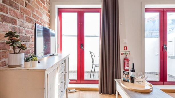 A modern kitchen with a brick wall, a flat-screen TV, a potted plant, and a red door leading outside. - GuestReady - Crayon Walls 3rd (Porto)