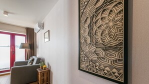A modern hotel room with a grey sofa, a small wooden side table with a lamp, and a large decorative wall panel. - GuestReady - Crayon Walls 3rd (Porto)