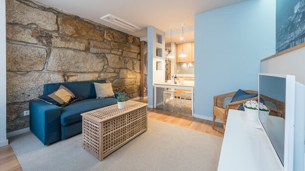 A modern living room with a stone wall, a blue sofa, a wooden coffee table, and a kitchen area in the background. - GuestReady - Crayon Walls Balcony (Porto)
