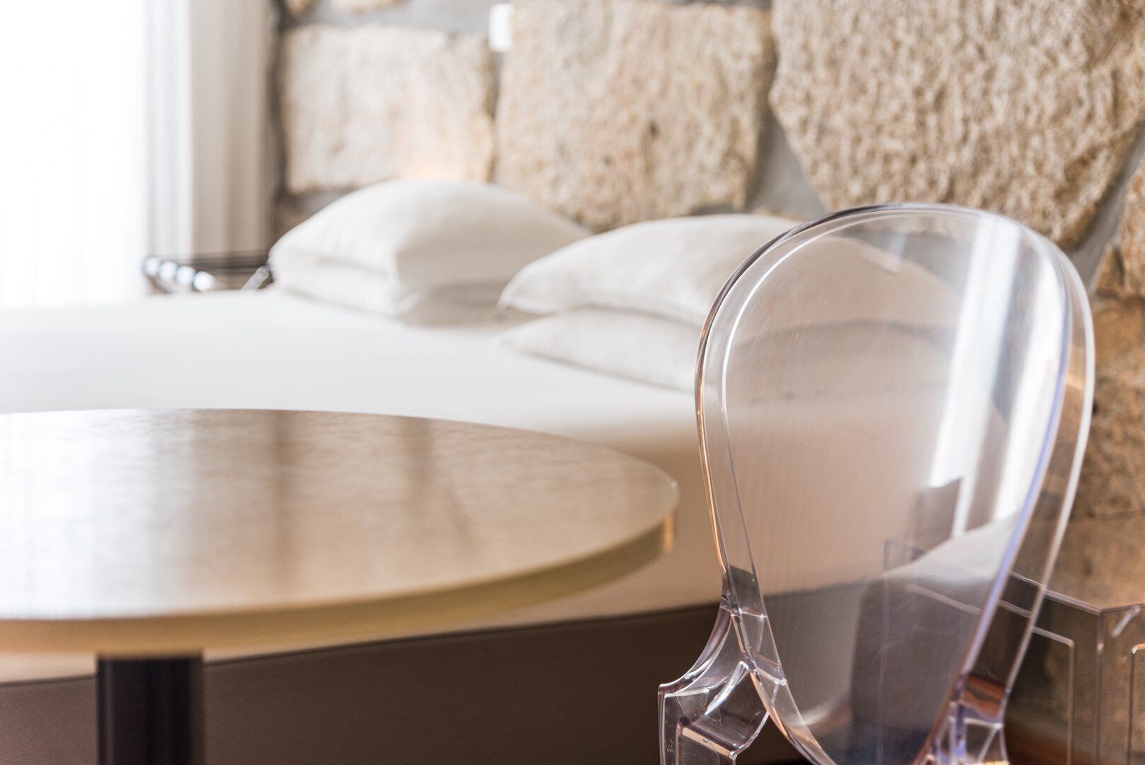 A modern hotel room with a round table, a clear chair, and a bed with white linens.
