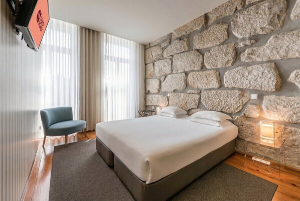 A hotel room with a stone wall, a bed with white bedding, a blue chair, and a TV mounted on the wall. - GuestReady - Porto de Rio 2.2 (Porto)