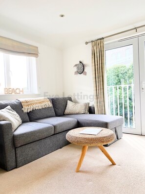 A modern living room with a grey sectional sofa, a woven ottoman, and a balcony with curtains. - The perfect seaside retreat - located next to Porthminster beach WITH parking (Saint Ives)