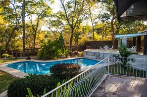A backyard with a swimming pool, surrounded by trees and a covered patio area with outdoor furniture. - Cosy Family Home in glen Lorne  (Harare)