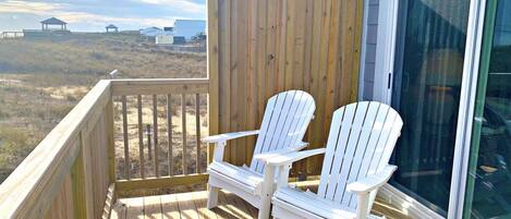 A wooden deck with two white Adirondack chairs, a glass door, and a view of a beach and buildings in the distance.
