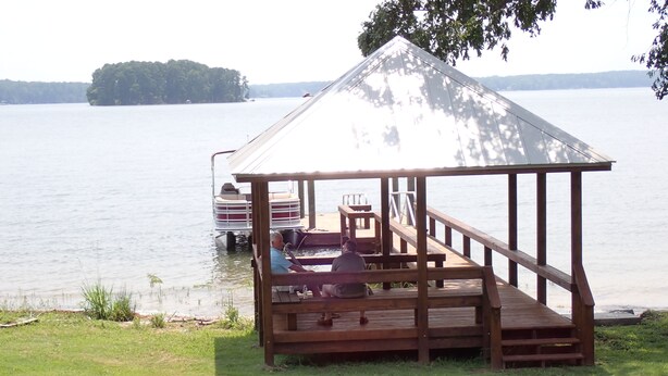 Une terrasse en bois avec un belvédère, un bateau amarré à proximité, et un lac avec une île qui se dessine à l’horizon.