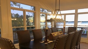 Un espace repas comprenant une grande table et six chaises, de grandes fenêtres offrant une vue sur des arbres et un plan d’eau, ainsi qu’un luminaire suspendu.