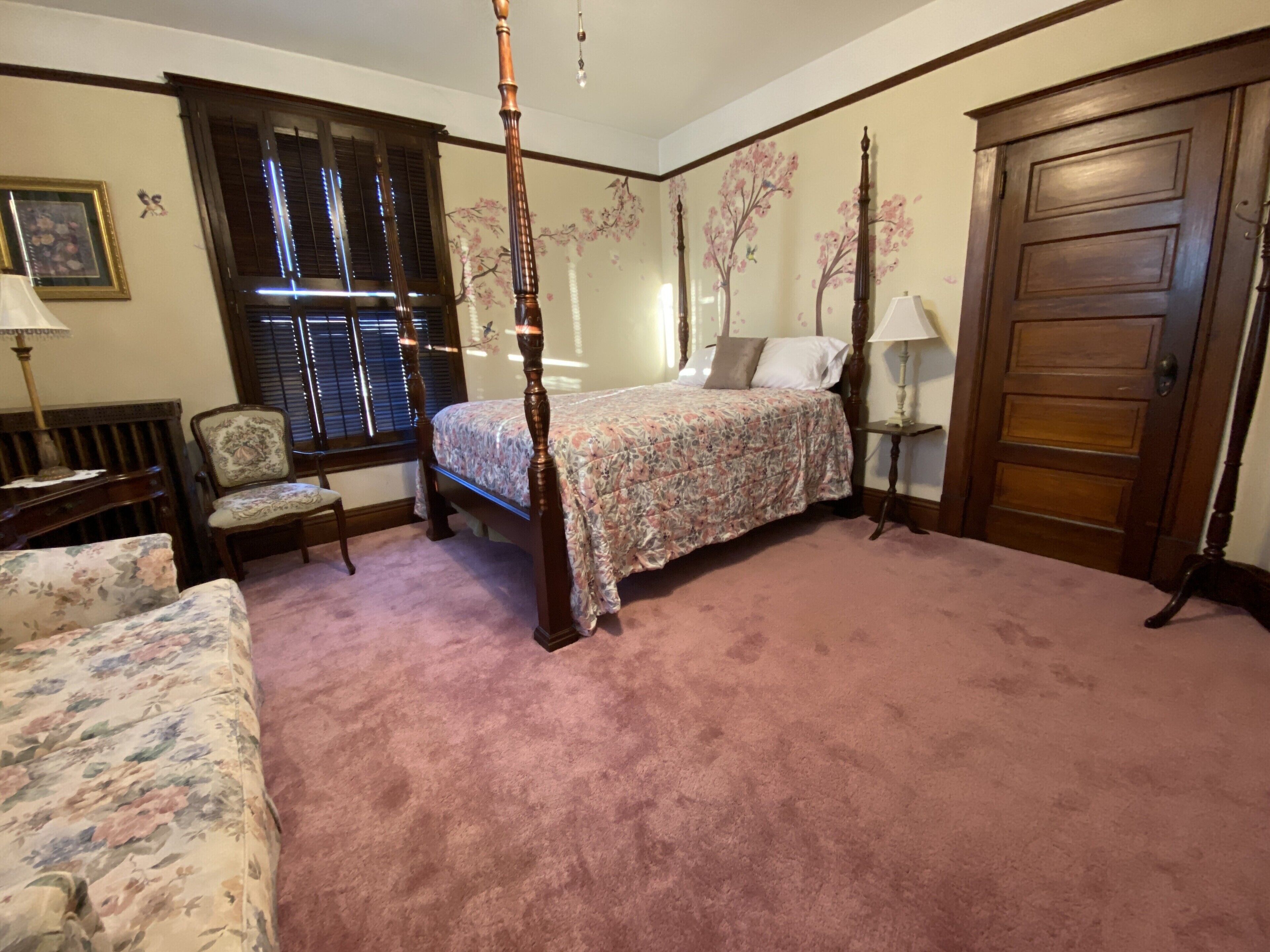 A bedroom with a four-poster bed, a floral-patterned sofa, a wooden chair, and a wooden door.