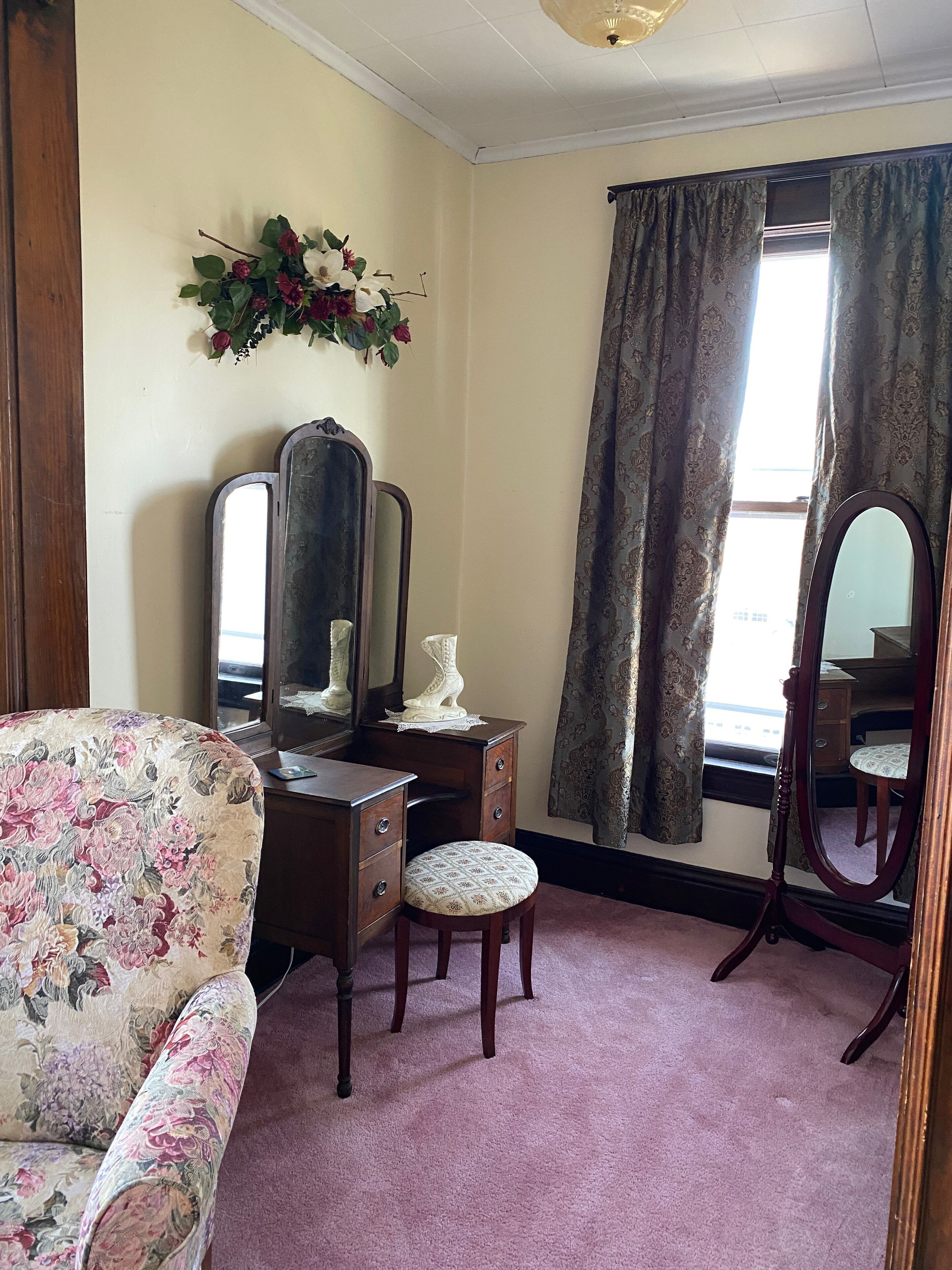 Ein Zimmer mit einem blumengemusterten Sofa, einem Holzschrank mit Spiegel, einem Schminktisch mit Spiegel und einem Fenster mit gemusterten Vorhängen.
