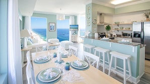 A dining area with a view of the ocean, a kitchen with a refrigerator, and a bar area with stools.
