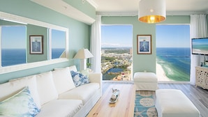 A spacious living room with a large window offering a view of the ocean and a cityscape. There is a white sofa, a coffee table, and a TV on a cabinet.