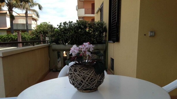 Une table ronde blanche, sur laquelle repose un panier tressé contenant des fleurs roses, est placée devant un mur jaune orné d’une balustrade de balcon.