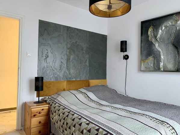 A bedroom with a bed, a wooden nightstand, a modern pendant light, and a large abstract painting on the wall.