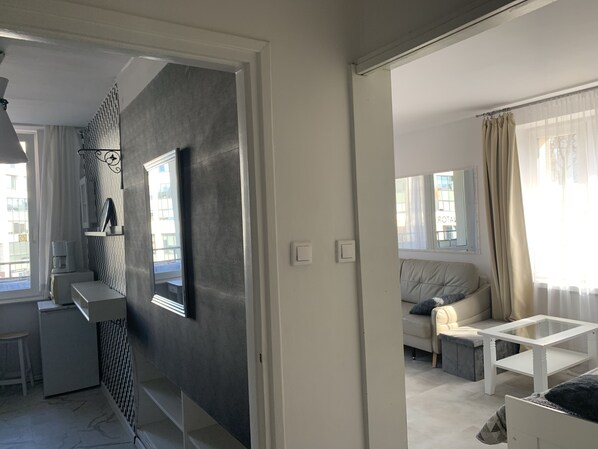 Un salón moderno con un sofá, una mesa de centro y una mesita auxiliar. Hay una ventana con cortinas, y se ve un radiador en la pared.