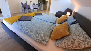 Une chambre moderne avec un grand lit, un petit canapé et un coin repas, avec un téléviseur et une cuisine en arrière-plan.