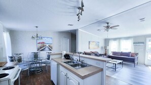 Eine moderne Küche mit Kochinsel, ein Essbereich mit Glastisch und ein Wohnzimmer mit Sofa und Deckenventilator.