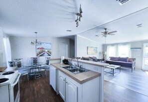 A modern kitchen with a central island, a dining area with a glass table, and a living room with a sofa and a ceiling fan.