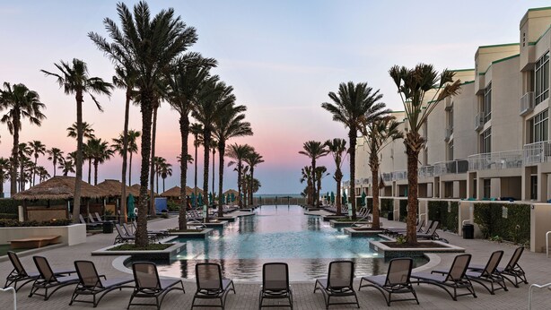 A resort pool area with lounge chairs, palm trees, and a building with balconies. - Life is beautiful at Sapphire on SPI! (South Padre Island)