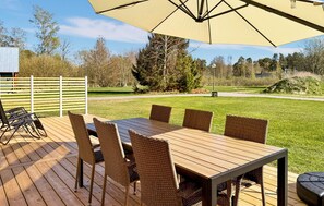 An outdoor patio with a wooden table and wicker chairs, an umbrella, and a view of a grassy area with trees.