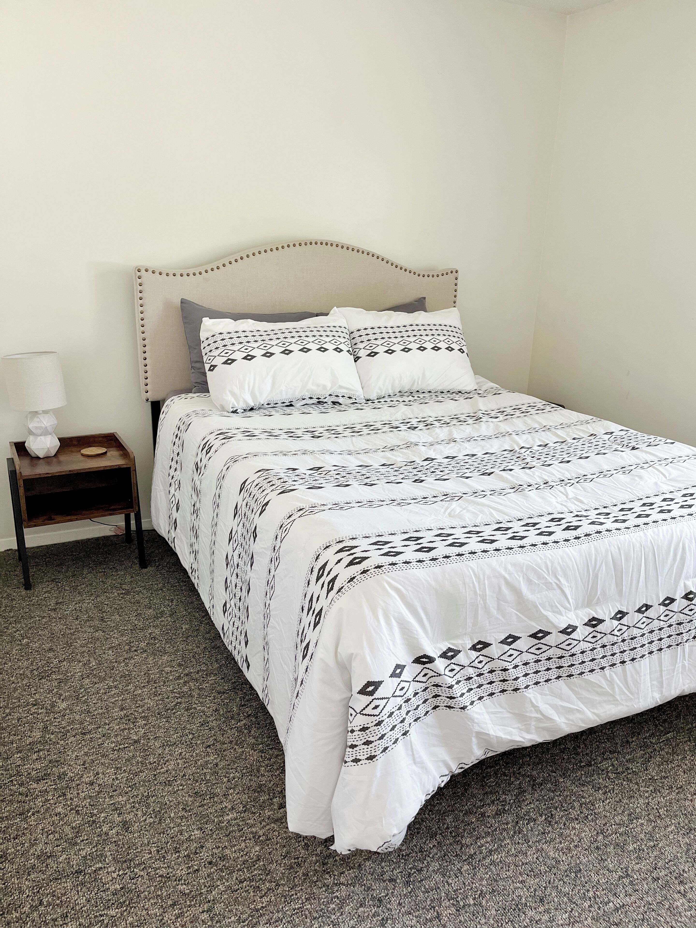 A neatly made bed with a patterned bedspread and pillows, a bedside table with a lamp, and a carpeted floor.